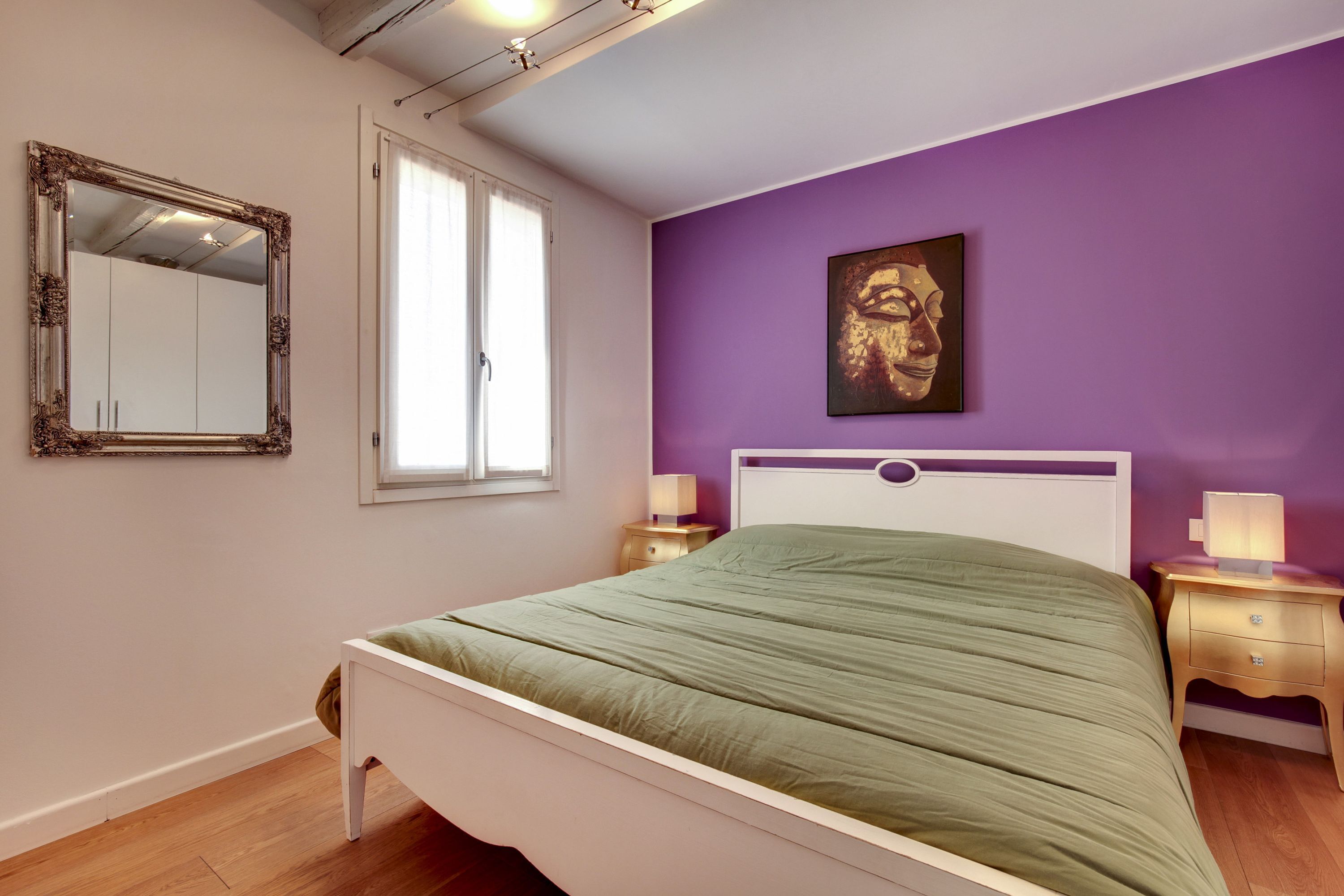 Ein Schlafzimmer mit einem Bett, einem Fenster, einem Spiegel, einem Nachttisch und einer Wand mit einem Gemälde.
