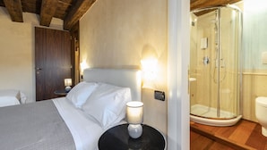 A hotel room with a bed, bedside lamp, wooden ceiling beams, and a glass-enclosed shower area. - Doge Palace 2 - Locz (Venice)