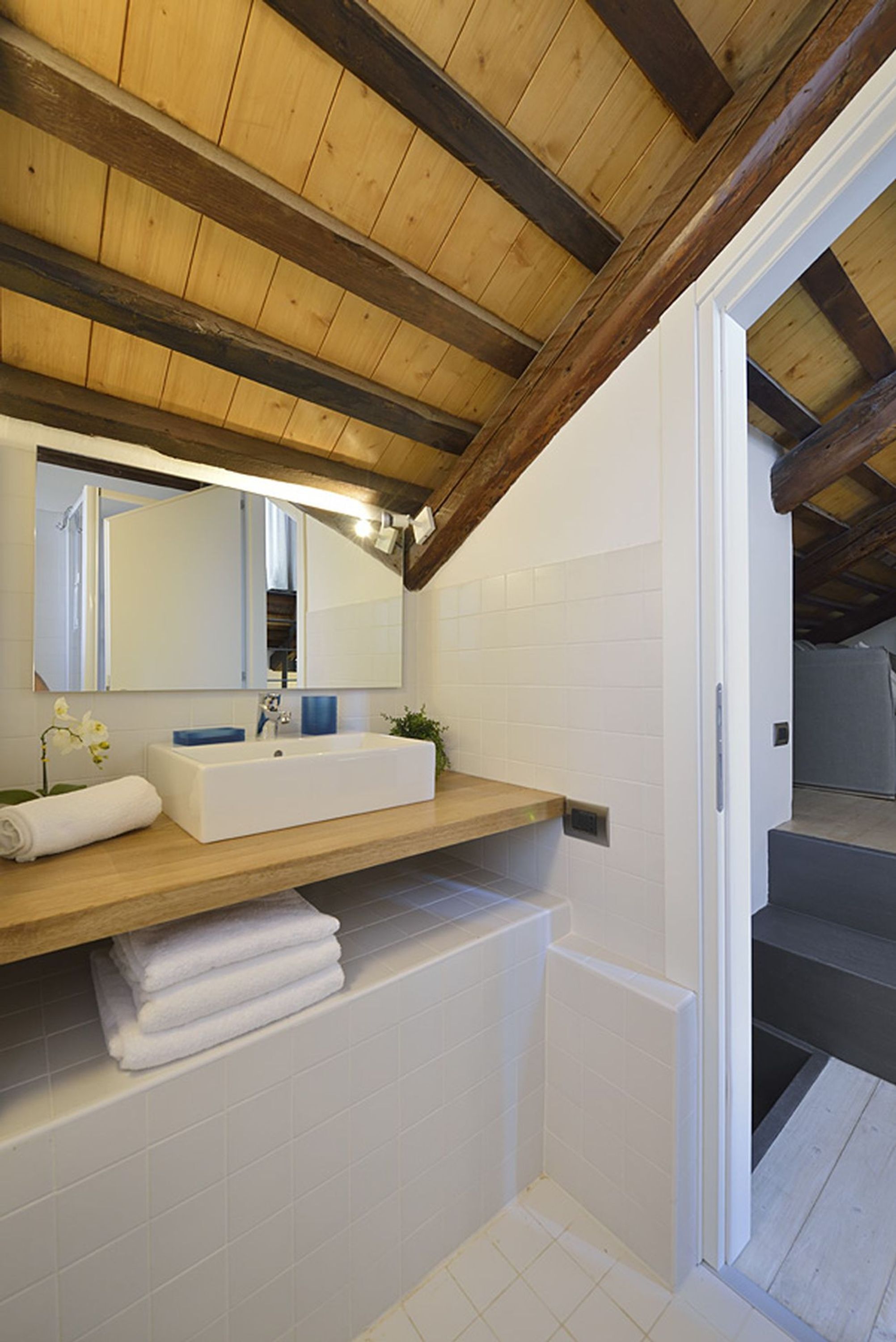 A modern bathroom with a wooden ceiling, white tiles, a large mirror, a white sink, and a towel rack.