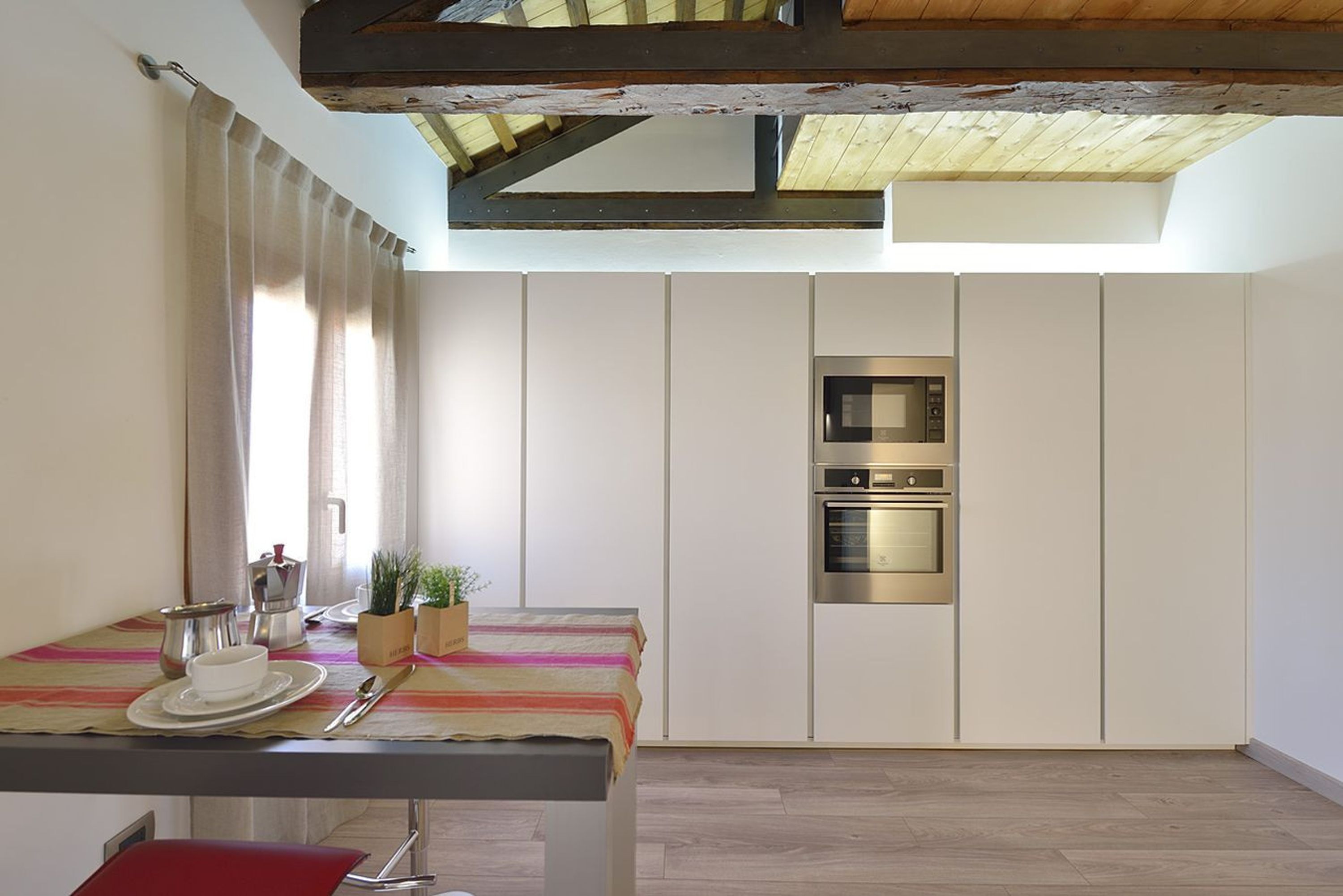 Een moderne keuken met een ingebouwde oven en magnetron, een eettafel met een geruit tafelkleed, en een rode stoel.