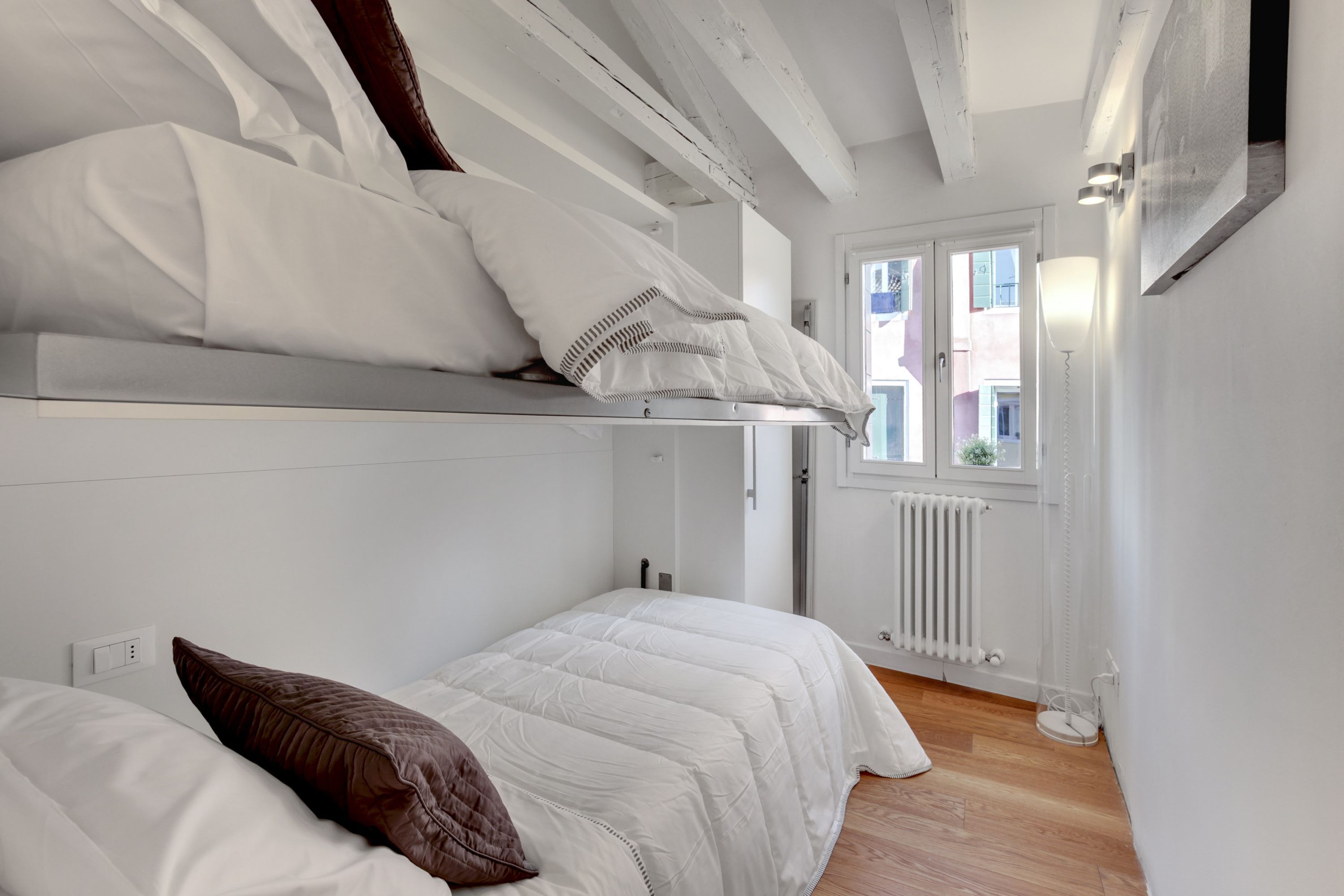 Ein Schlafraum mit zwei Betten, weißer Bettwäsche, Holzböden, einem Heizkörper und einem Fenster mit Blick auf Gebäude.