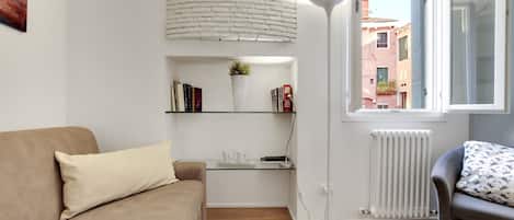 A living room with a beige sofa, a glass coffee table, a white floor lamp, and a view of a cityscape through a window.