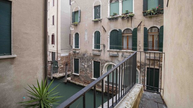 Un étroit canal vénitien bordé de bâtiments des deux côtés, avec des fenêtres en arc et des balcons ornés de plantes.