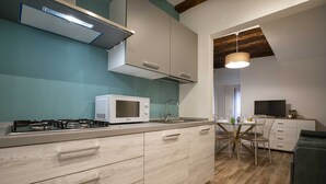 Une cuisine moderne équipée d’une cuisinière à gaz, d’un four à micro-ondes et de meubles blancs. On y trouve un coin repas avec une table et des chaises, ainsi qu’un téléviseur posé sur un meuble.