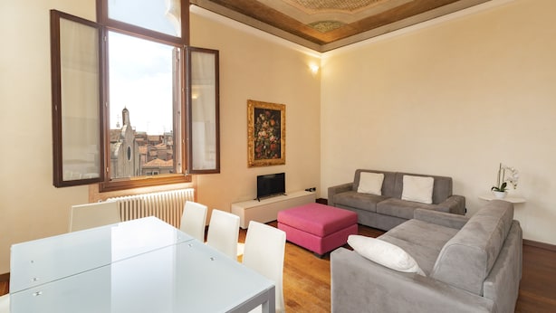 A modern living room with a grey sofa, white chairs, a glass table, and a pink ottoman. A painting is on the wall, and a window offers a city view. - Doge Palace 7 - Locz (Venice)