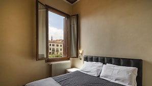 A hotel room with a bed, a window with curtains, a radiator, and a view of buildings outside. - Doge Palace 7 - Locz (Venice)