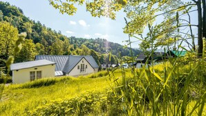A house with a sloped roof and a small porch, surrounded by lush greenery and hills in the background. - Villa met Sauna Ldd032 (Desna)