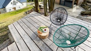 A wooden deck with two chairs, a small table with flowers and a cup, and a view of a house and trees in the background. - Villa met Sauna Ldd032 (Desna)