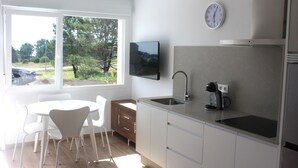 A modern kitchen with white cabinets, a built-in sink, and a coffee maker. There is a dining table with chairs and a window with a view of trees. - Alén de Louro - Apartamento 1 San Francisco (Lariño)