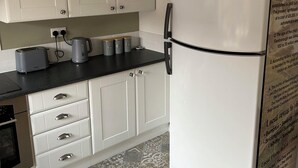 Eine Küche mit weißen Schränken, einer schwarzen Arbeitsplatte und einem weißen Kühlschrank. Auf der Arbeitsplatte befinden sich zwei Wasserkocher, ein Toaster und ein weiterer Wasserkocher. Der Boden ist mit einem gemusterten Fliesenbelag versehen.