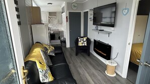 Kompakti oleskelutila, jossa on sohva, televisio, takka ja avoimen oven kautta näkyvä keittiö.