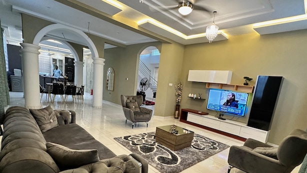 Uma sala de estar espaçosa com um amplo sofá em formato de L, uma poltrona, uma mesa de centro e uma televisão de tela plana fixada na parede.