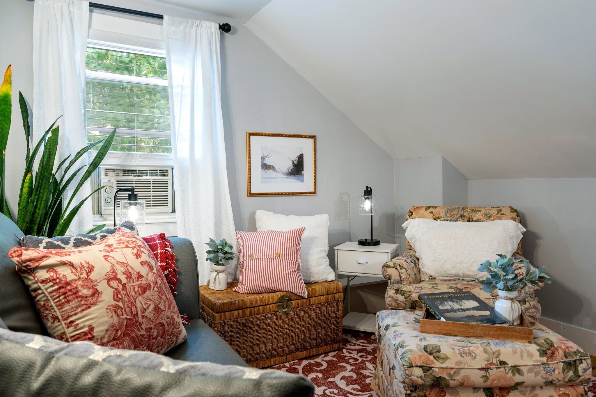 Una sala de estar acogedora con un sofá, un cofre de mimbre, un sillón estampado y una mesita de noche con lámpara.