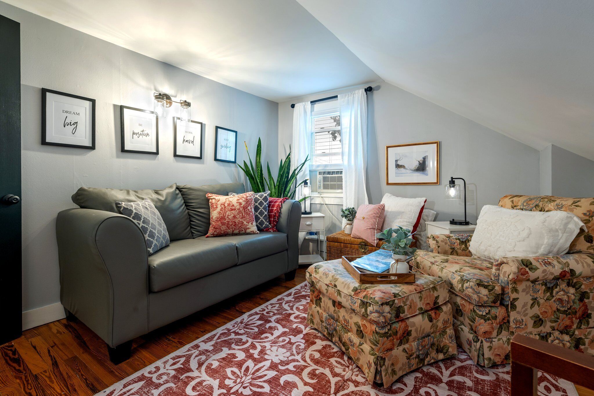 A living room with a grey sofa, a floral armchair, and a patterned rug. There are framed pictures on the wall and a plant on the floor.