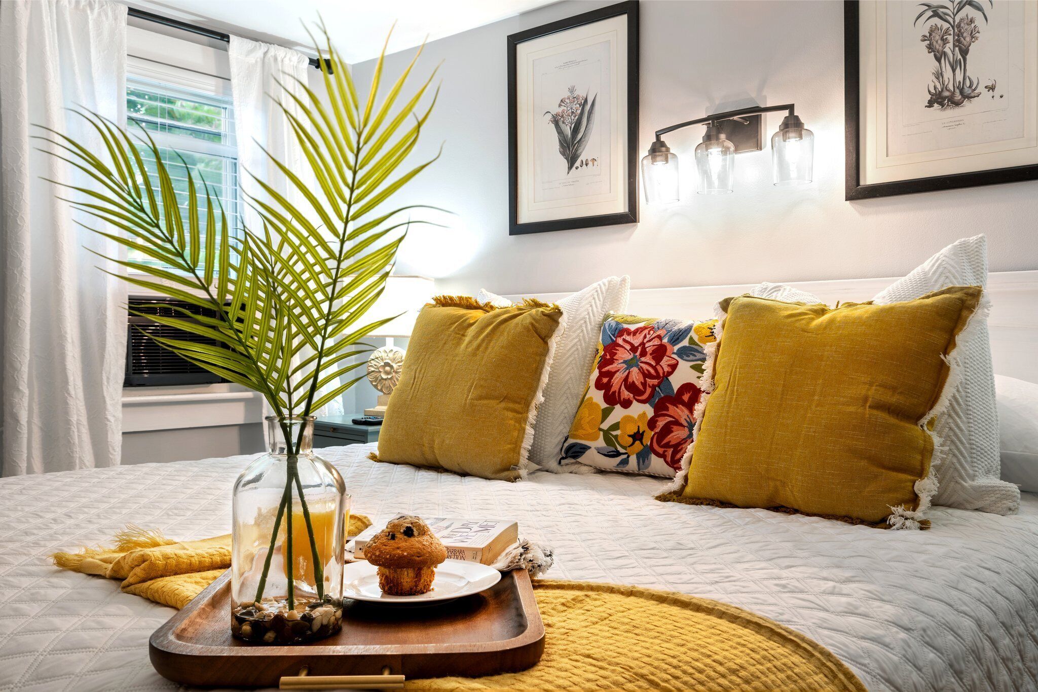 Een netjes opgemaakt bed met een dienblad vol ontbijtartikelen, een potplant en een ingelijste botanische afbeelding aan de muur.