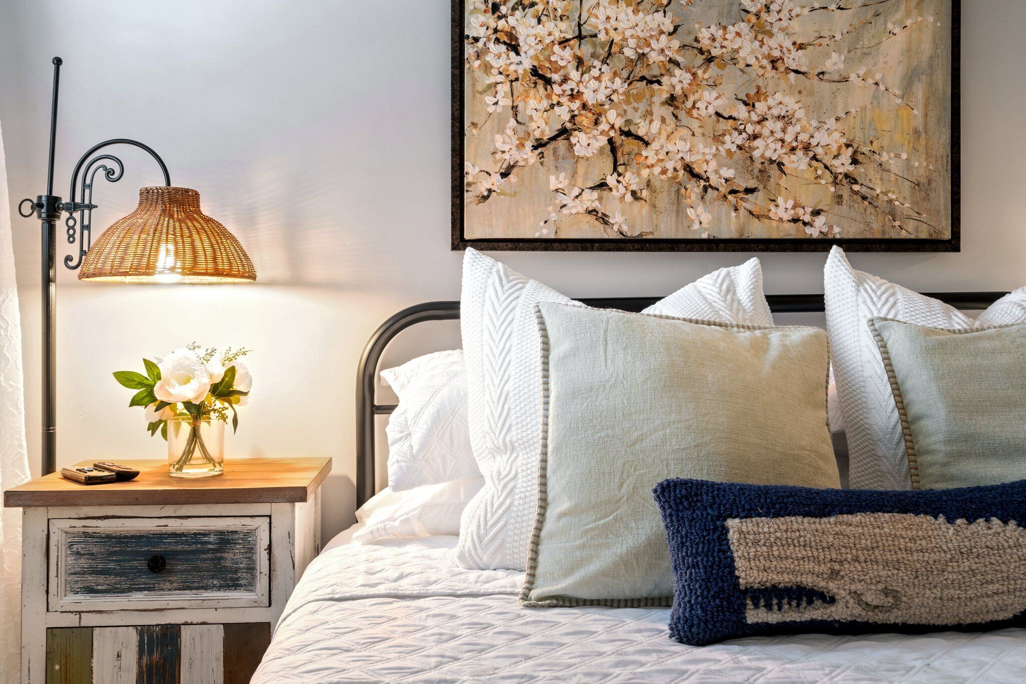 Cama bem arrumada com almofadas brancas e azuis, um abajur e um vaso com flores sobre uma mesa de cabeceira de madeira.