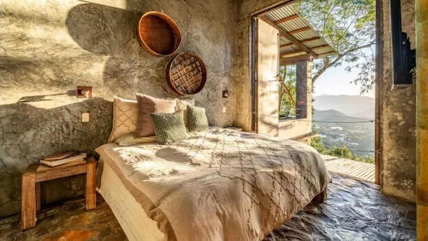 Un dormitorio con pared de piedra, una cama con colcha estampada, una mesita de noche de madera y vistas al paisaje a través de un vano abierto.