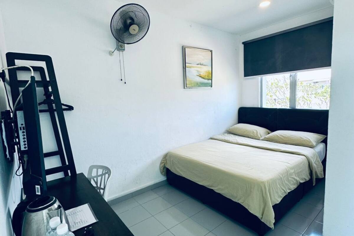 Un dormitorio con una cama, un escritorio con hervidor, una botella de agua y un ventilador.