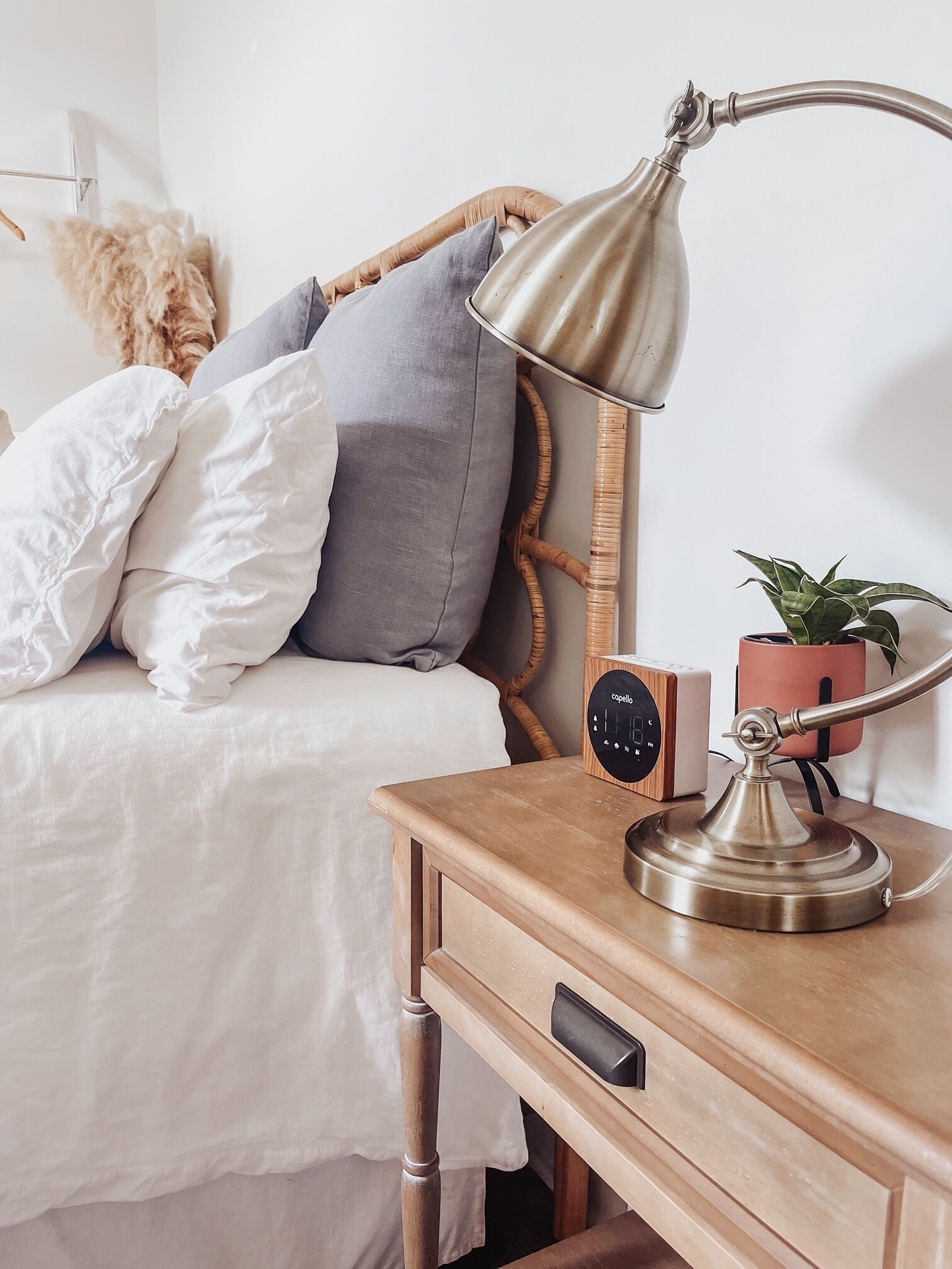 Un lit bien fait, avec une literie blanche et grise, une table de chevet en bois sur laquelle se trouve une lampe, un petit haut-parleur et une plante en pot.