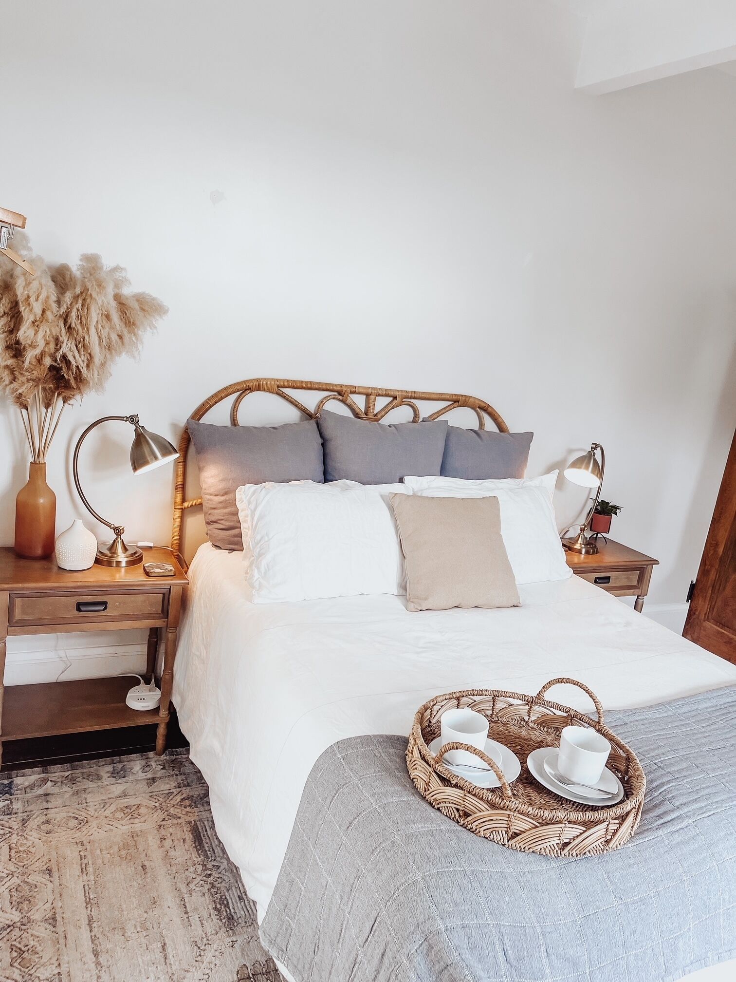 Een netjes opgemaakt bed met een grijze deken, een rieten dienblad met kopjes en schoteltjes, en een nachtkastje met een lamp en een vaas gedroogde bloemen.
