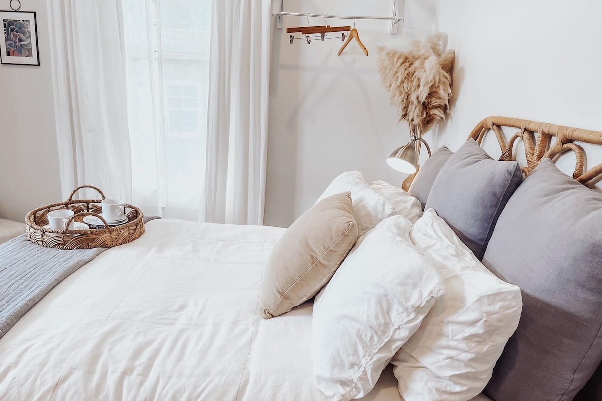 A neatly made bed with white bedding and multiple pillows, a wicker tray with cups and saucers, and a hanging plant with dried flowers.