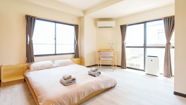 A spacious hotel room with a large bed, two windows with curtains, a small desk with a chair, and a balcony door.