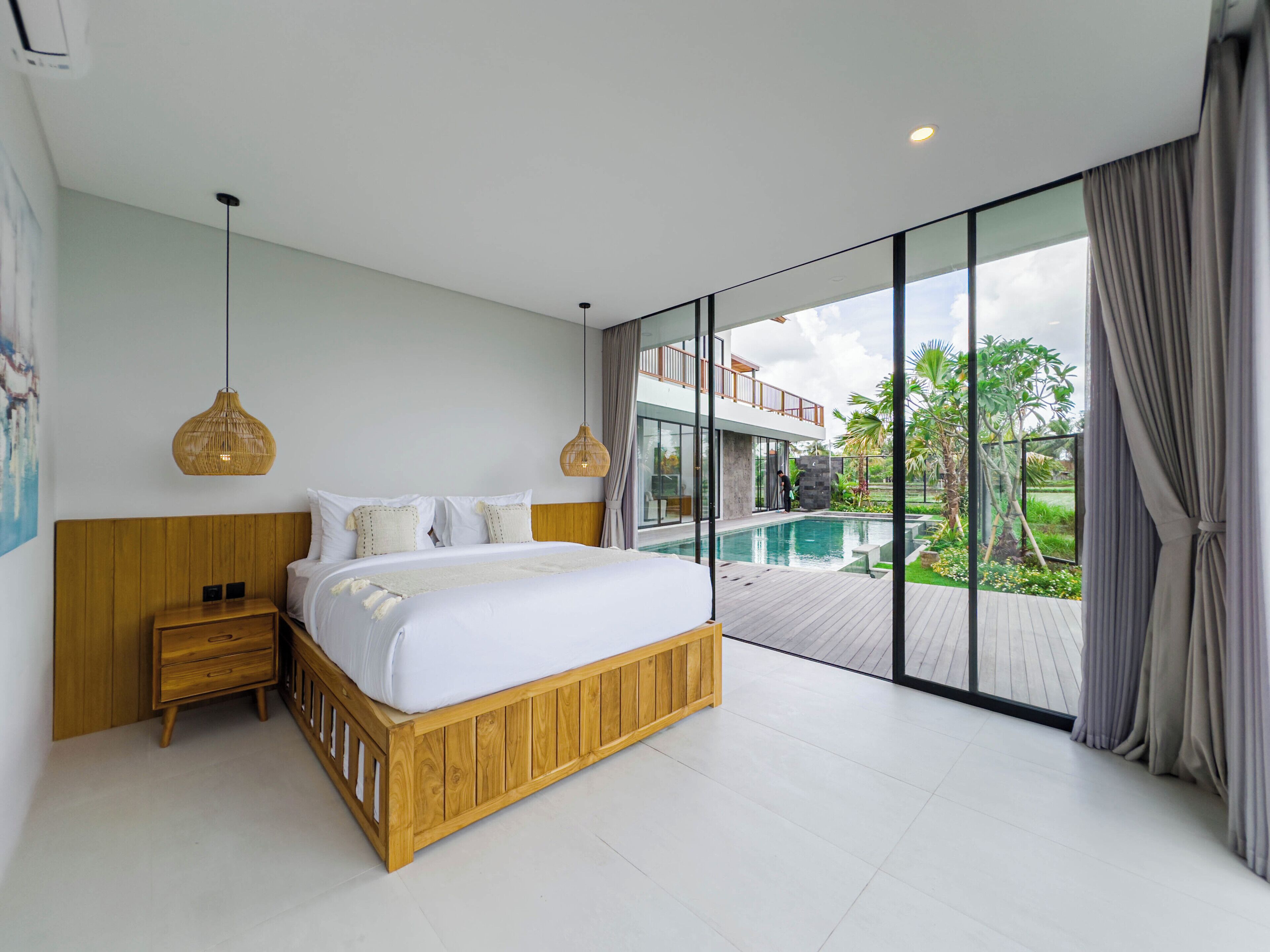 A modern bedroom with a large bed, wooden headboard, bedside tables, and a view of a swimming pool and outdoor area.