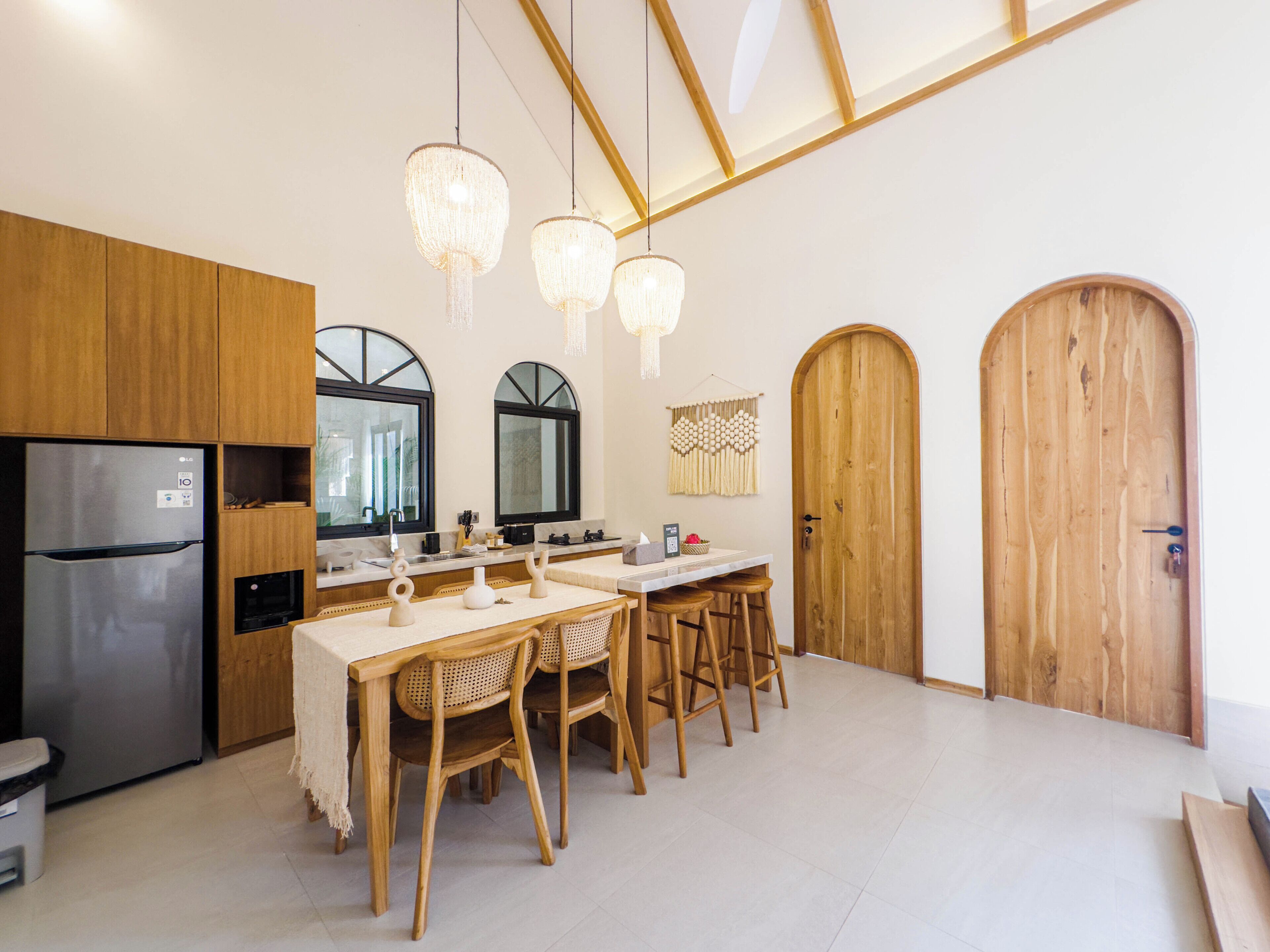 A modern kitchen with wooden cabinets, a stainless steel refrigerator, a dining area with a table and chairs, and a wooden door.