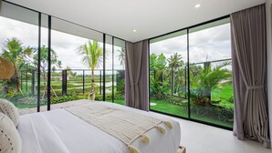 A bedroom with a large bed, a view of a lush garden through floor-to-ceiling windows, and a balcony with a view of the garden.