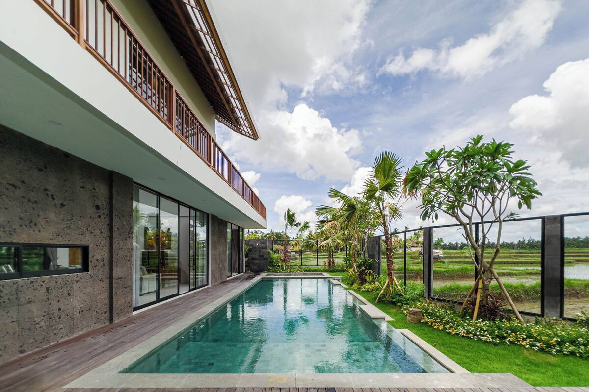 A modern house with a swimming pool, large windows, and a balcony overlooking a garden and water body.