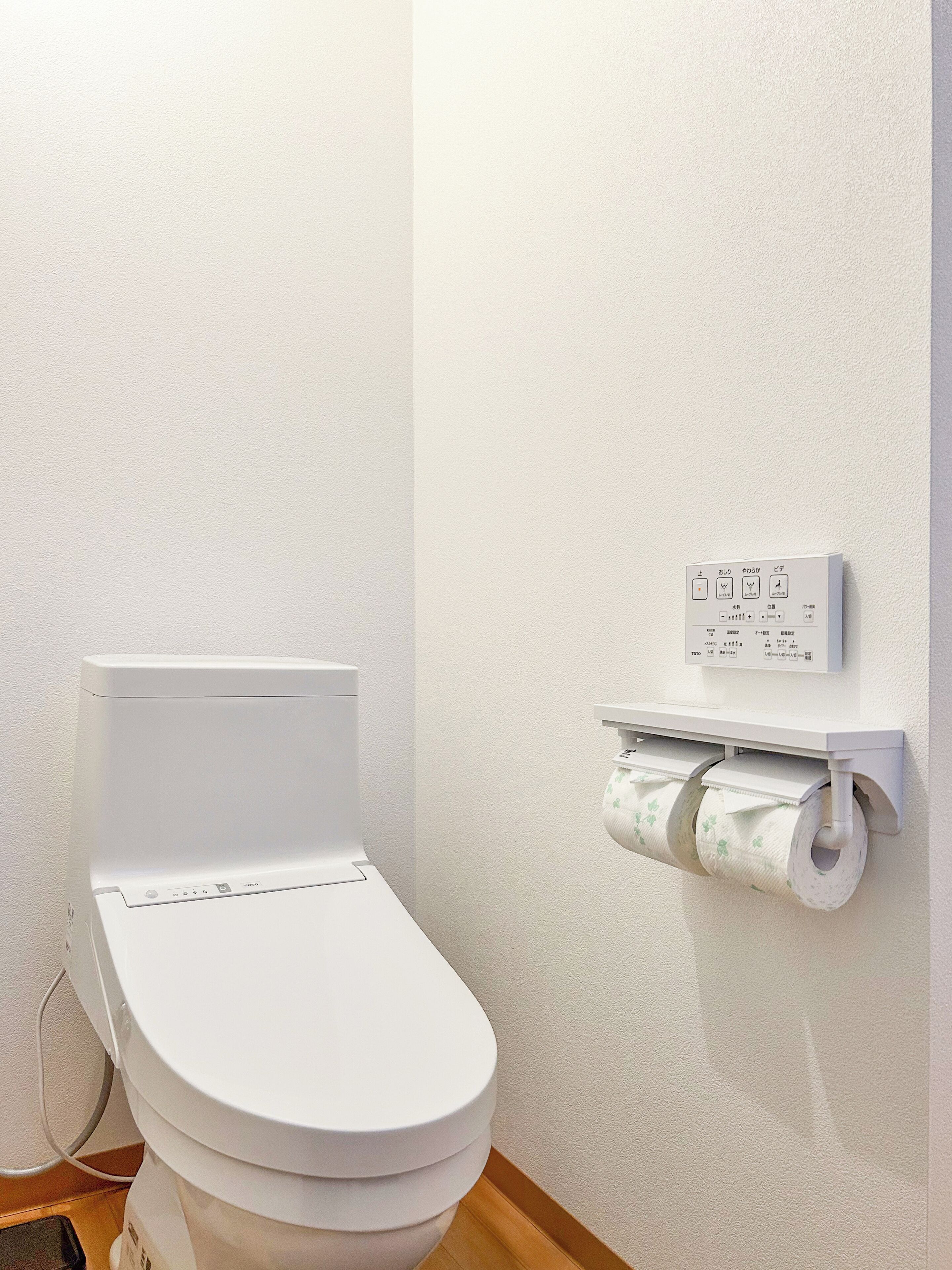 A white toilet with a control panel on the wall, a toilet paper holder, and a visible toilet paper roll.
