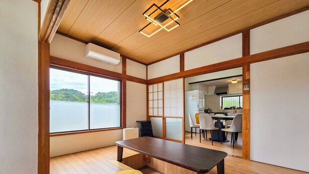 Una habitación con piso de madera, una mesa de comedor, un reposapiés amarillo, vista a un lago y una cocina visible a través de una puerta abierta.