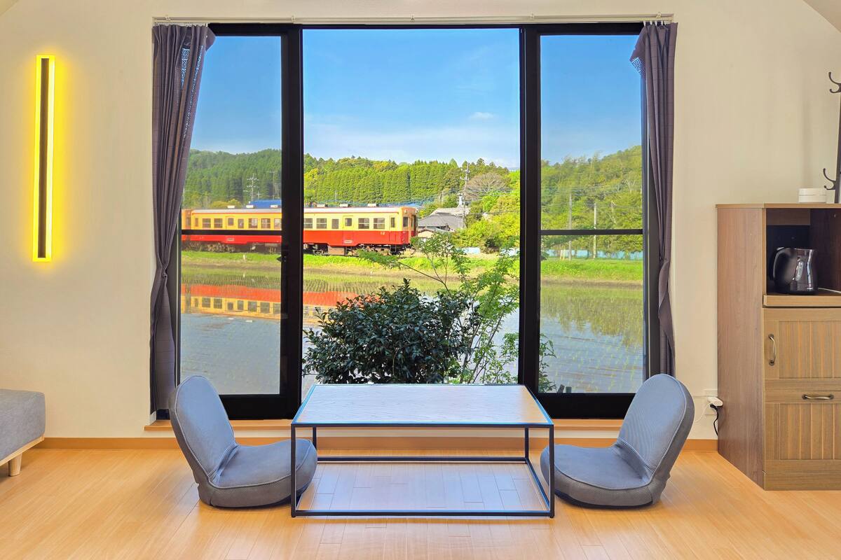 Ein Zimmer mit großem Fenster, einem Tisch, zwei Stühlen, einem Schrank und einer Wandleuchte.