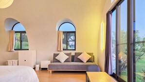 Un dormitorio con cama, sofá, una mesita y dos ventanas con cortinas.