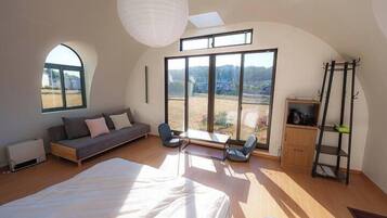 A spacious bedroom with a large bed, a sofa, and a dining table. There are large windows offering a view of the outdoors.