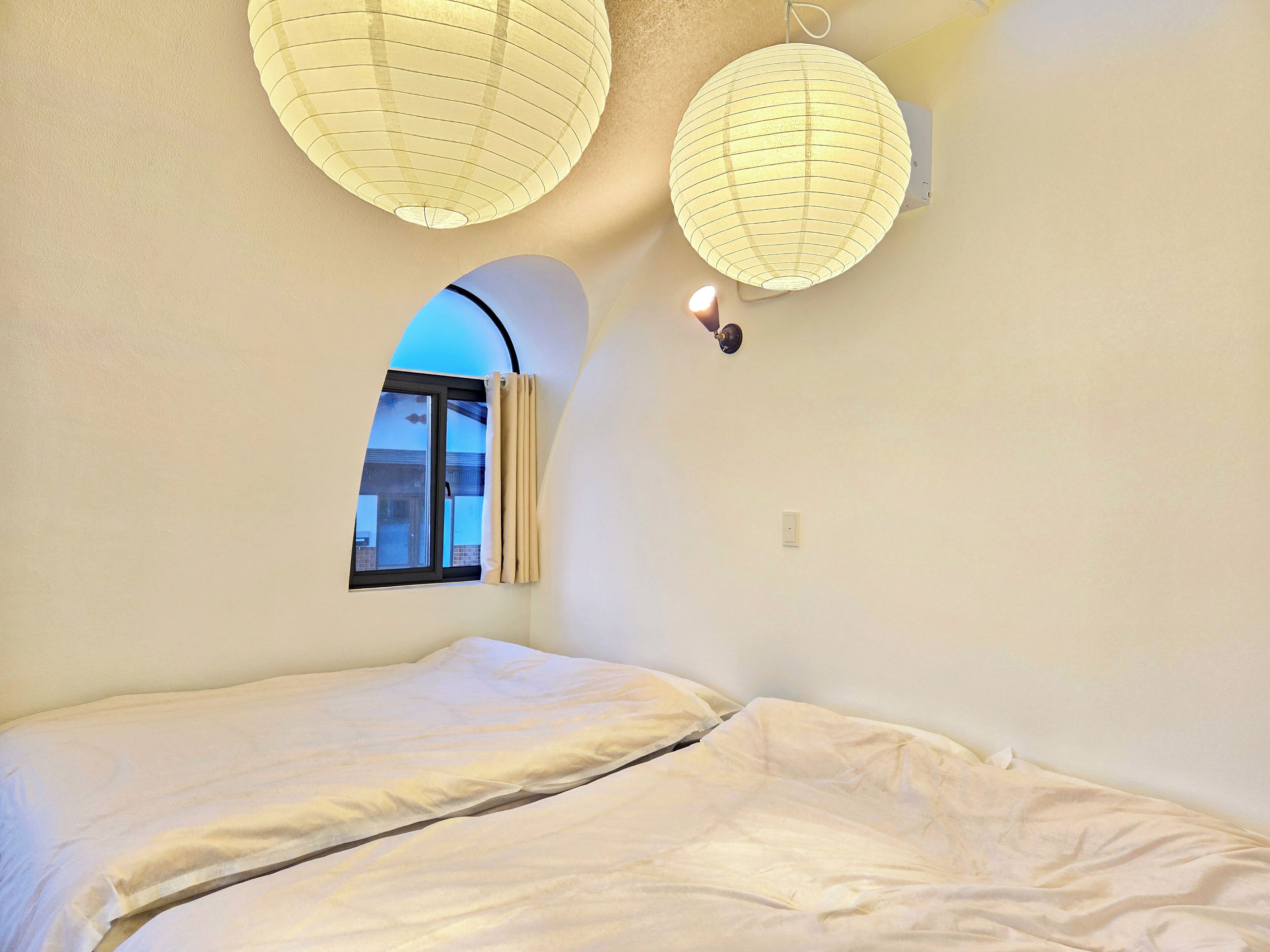 A bedroom with a bed, two hanging paper lanterns, a window with curtains, and a wall-mounted light fixture.