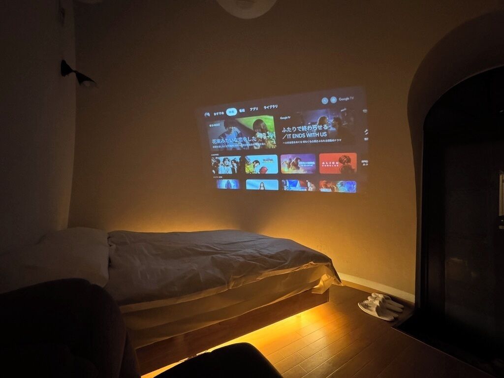A room with a bed, a wall-mounted projector displaying a video, and a wooden floor.