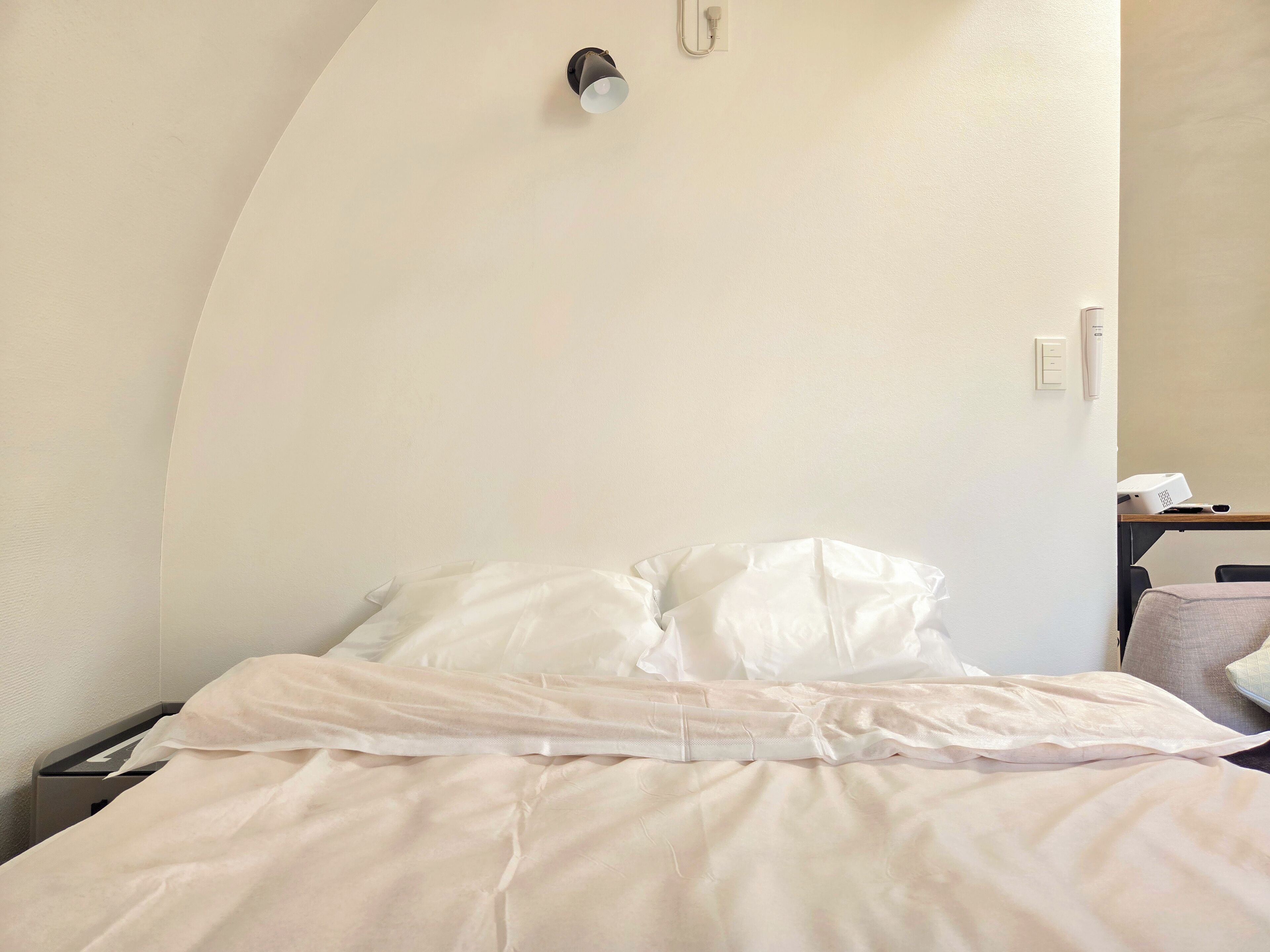 Una habitación de hotel con techo curvo, una cama con ropa de cama blanca, una mesita de noche y un pequeño escritorio con una lámpara y un dispositivo.