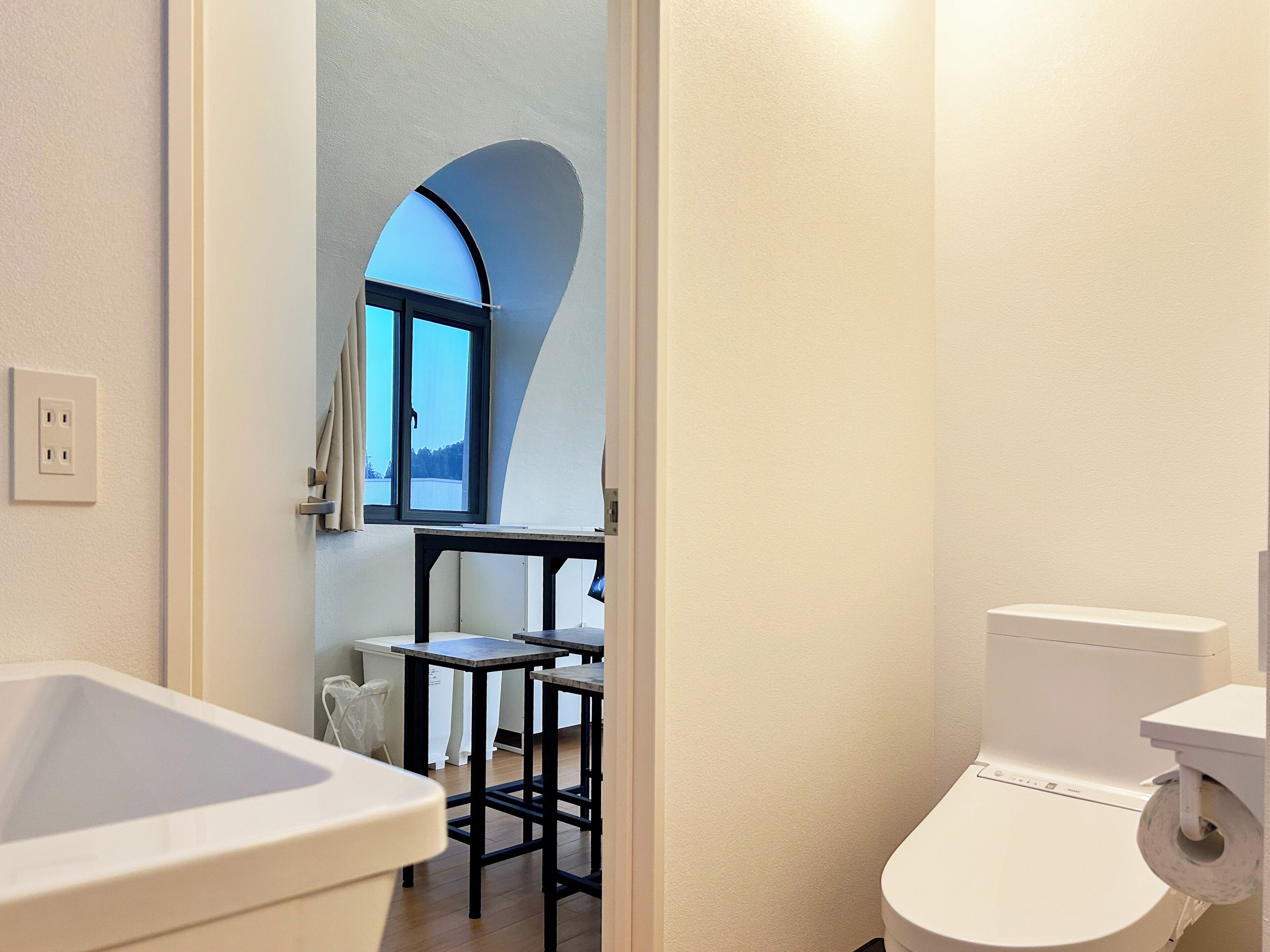 A modern bathroom with a white sink, a toilet, and a small table with a chair. There is a window with a view of the outdoors.