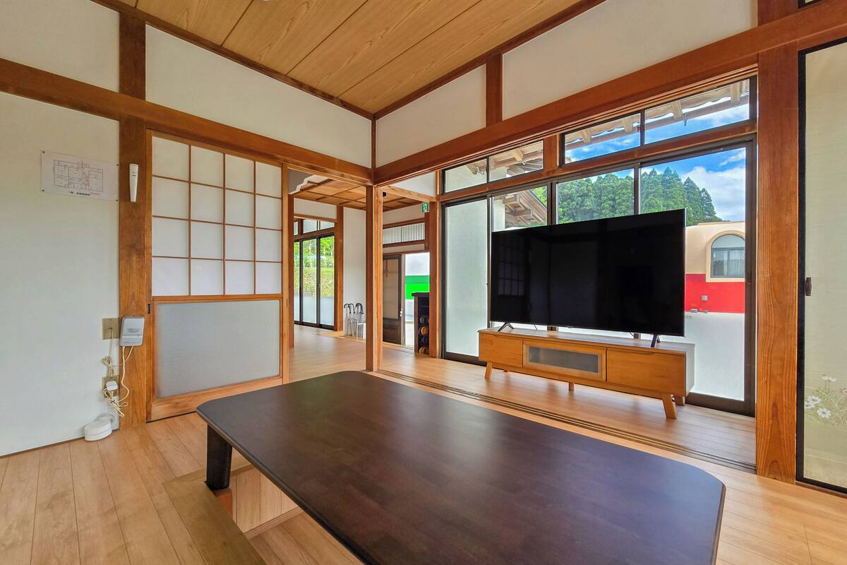 Ein geräumiges Zimmer mit einem großen, dunklen Holztisch, Glastüren, die auf einen Balkon führen, und einem Fernseher auf einem Holzständer.