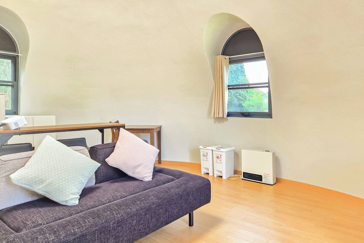 Ein modernes Wohnzimmer mit einem dunkelgrauen Sofa, zwei Kissen, einem Holztisch und einem Fenster mit Vorhängen. Auf dem Boden stehen zwei weiße Geräte.