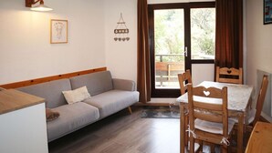 A cozy living room with a sofa, wooden dining table, and chairs. There is a window with curtains, a wall-mounted light, and a small wall decoration. - Nice studio 6 persons COEUR DE STATION (LES ORRES 1650)