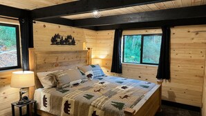 Camera da letto in una baita di legno, con un letto, un comodino, una lampada e una finestra vista alberi.