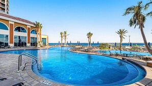 Una piscina grande y curvada con indicador de profundidad de 1.5 metros, rodeada de sillas de descanso y palmeras, con un edificio de grandes ventanales y techo de tejas al fondo.