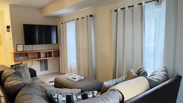 A modern living room with a large dark-colored sofa, a flat-screen TV mounted on the wall, and a window with curtains. -  Charming 3 bedroom home Centrally located in Worcester (Worcester)