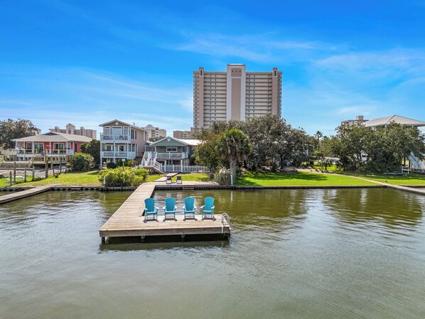 A waterfront property with a dock, lounge chairs, and a large building in the background. - Serenity Shores – 1 Block from Beach, Pet-Friendly! (Gulf Shores)