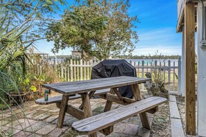 A wooden picnic table with benches in a backyard with a white fence, potted plants, and a view of a body of water. - Serenity Shores – 1 Block from Beach, Pet-Friendly! (Gulf Shores)