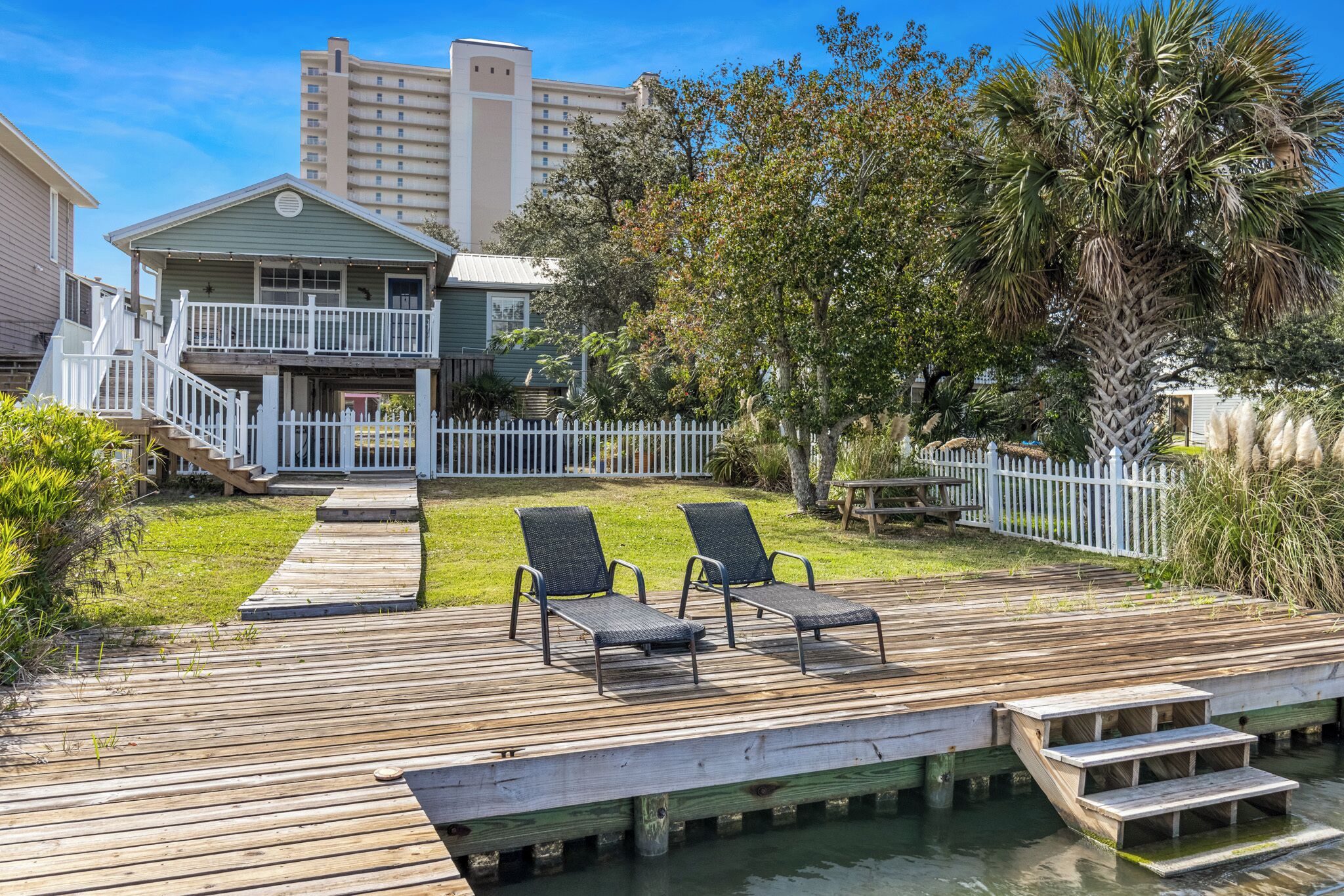 A wooden deck with two lounge chairs, a small pool, and a white fence surrounding a house with a balcony.
