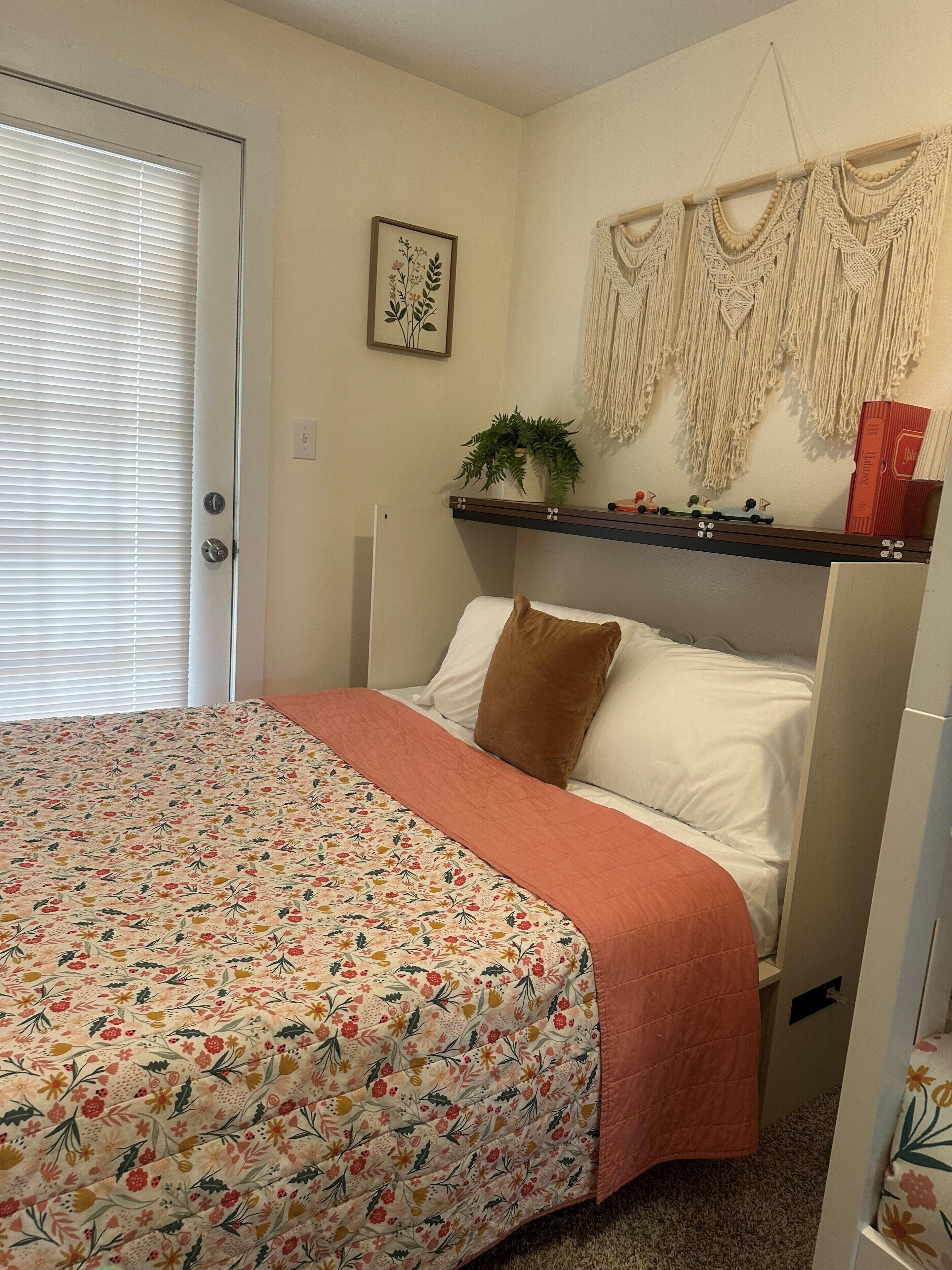 Une chambre avec une couvre-lit à motifs floraux, un oreiller marron, une étagère avec une plante et des livres, et une décoration murale en macramé.