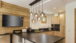 A modern kitchen with a large island, wooden cabinetry, a flat-screen TV, pendant lights, and a coffee machine. - Modern 2BR | MtBlanc View | Terrace | Brevent 3mn (Chamonix-Mont-Blanc)